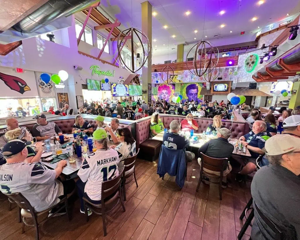 Bright, lively indoor sports bar filled with fans in jerseys, colorful wall art, and multiple screens showing sporting events.